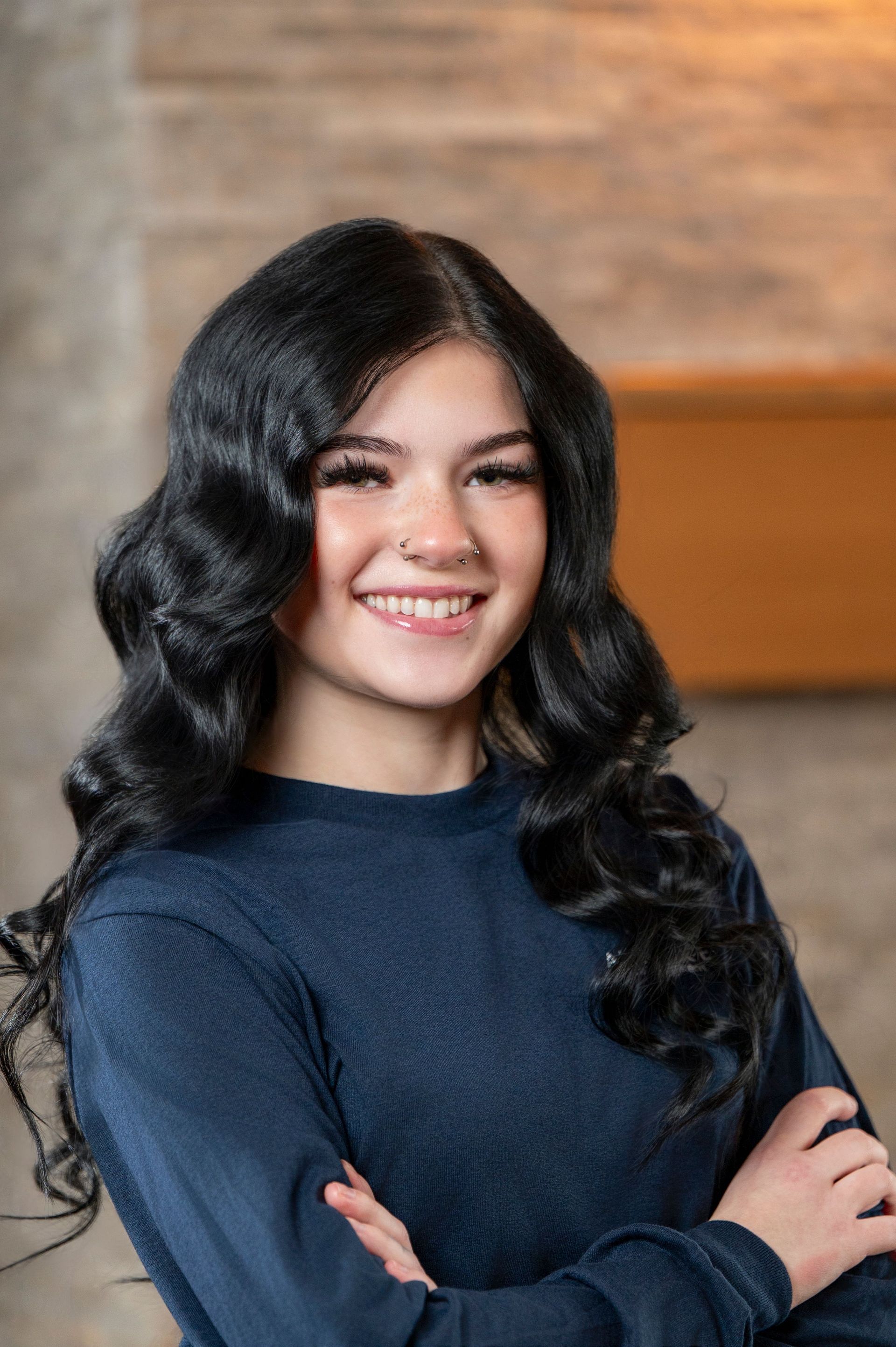 Smiling person with long black hair and crossed arms, wearing a navy sweater against a warm indoor background