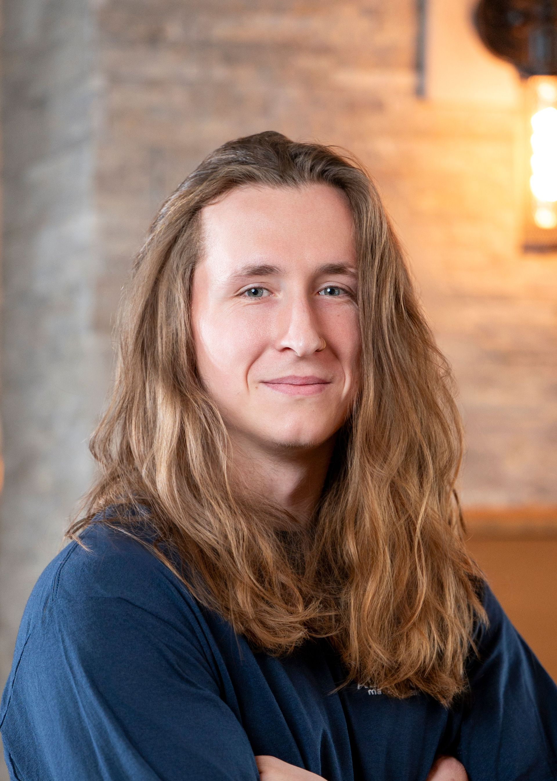 Portrait of a person with long hair in a dark blue shirt, seated indoors against a warm brick wall.