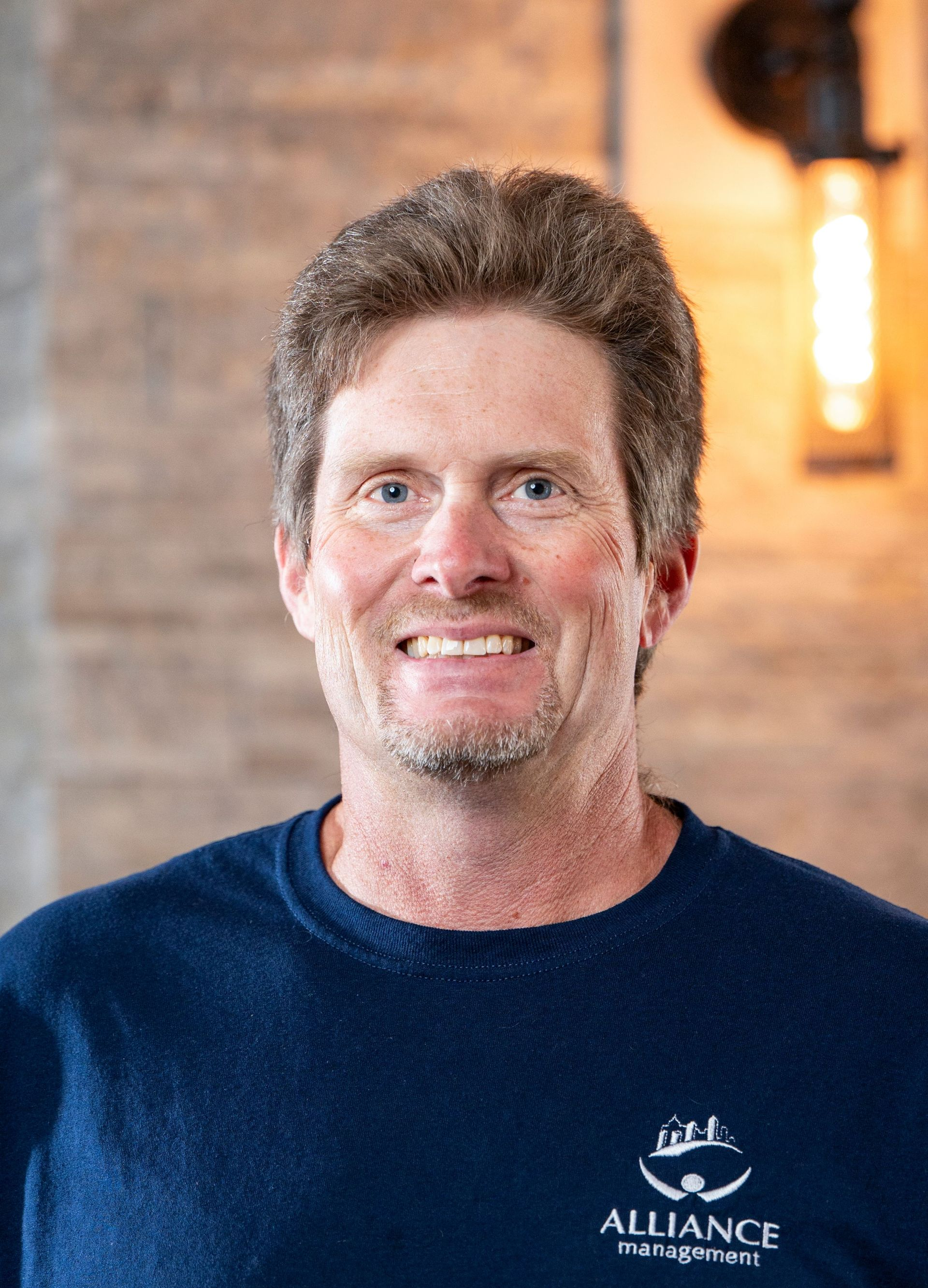Smiling man in a navy shirt with an Alliance logo, standing indoors against a warm brick wall.
