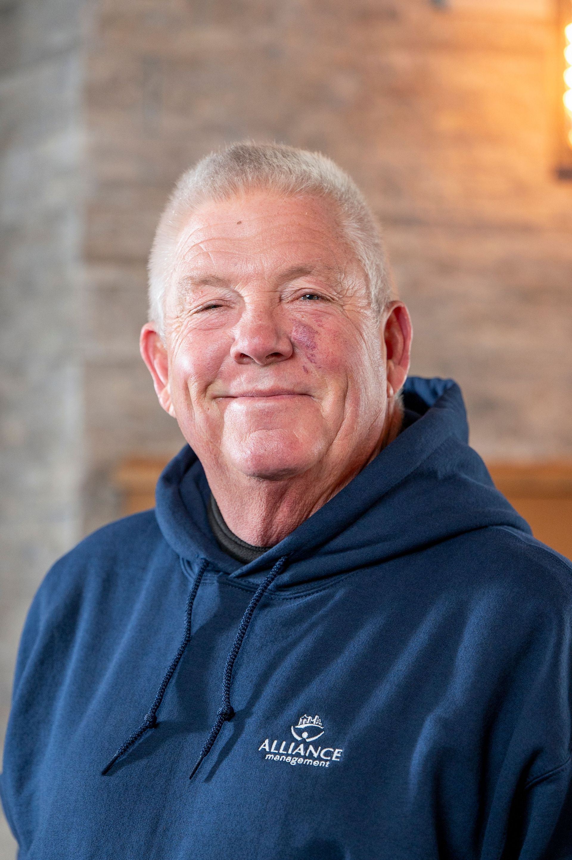 Smiling man in a navy hoodie standing indoors against a softly lit brick wall.