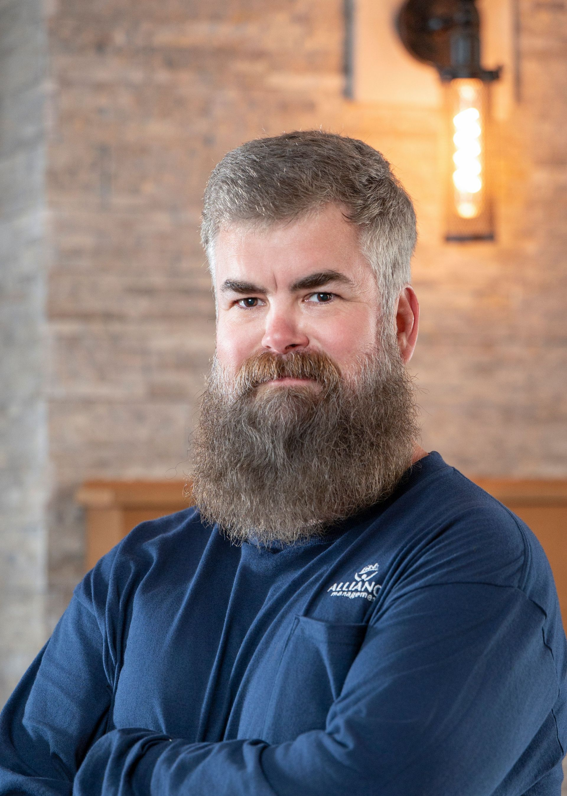 Bearded man in a blue shirt with arms crossed, standing indoors against a brick wall.