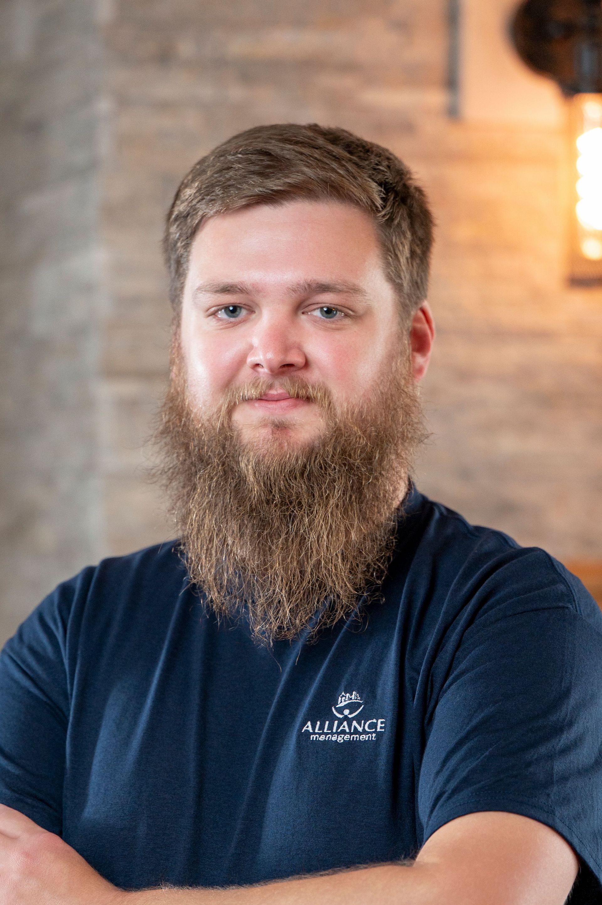 Bearded man in a navy T-shirt standing indoors against a blurred brick wall background