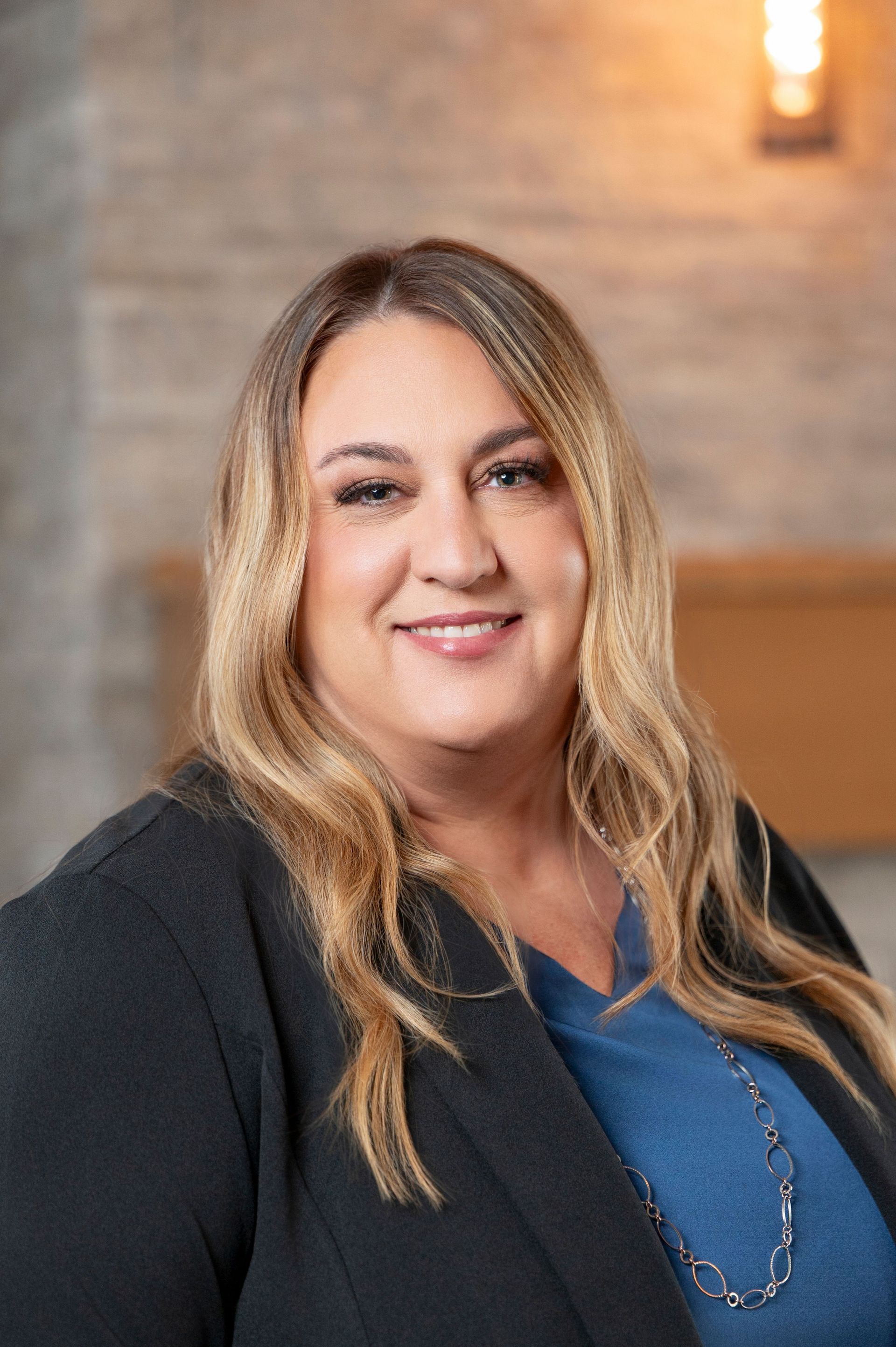 Smiling woman in a black blazer and blue top against a softly lit indoor background
