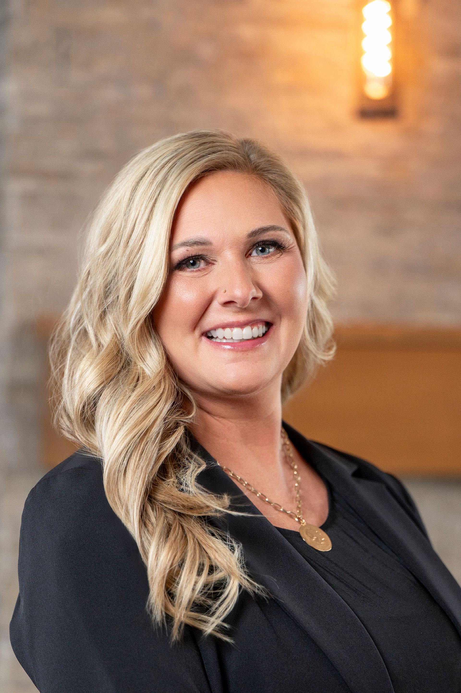 Smiling blonde woman in a black top, standing indoors against a softly lit brick wall.