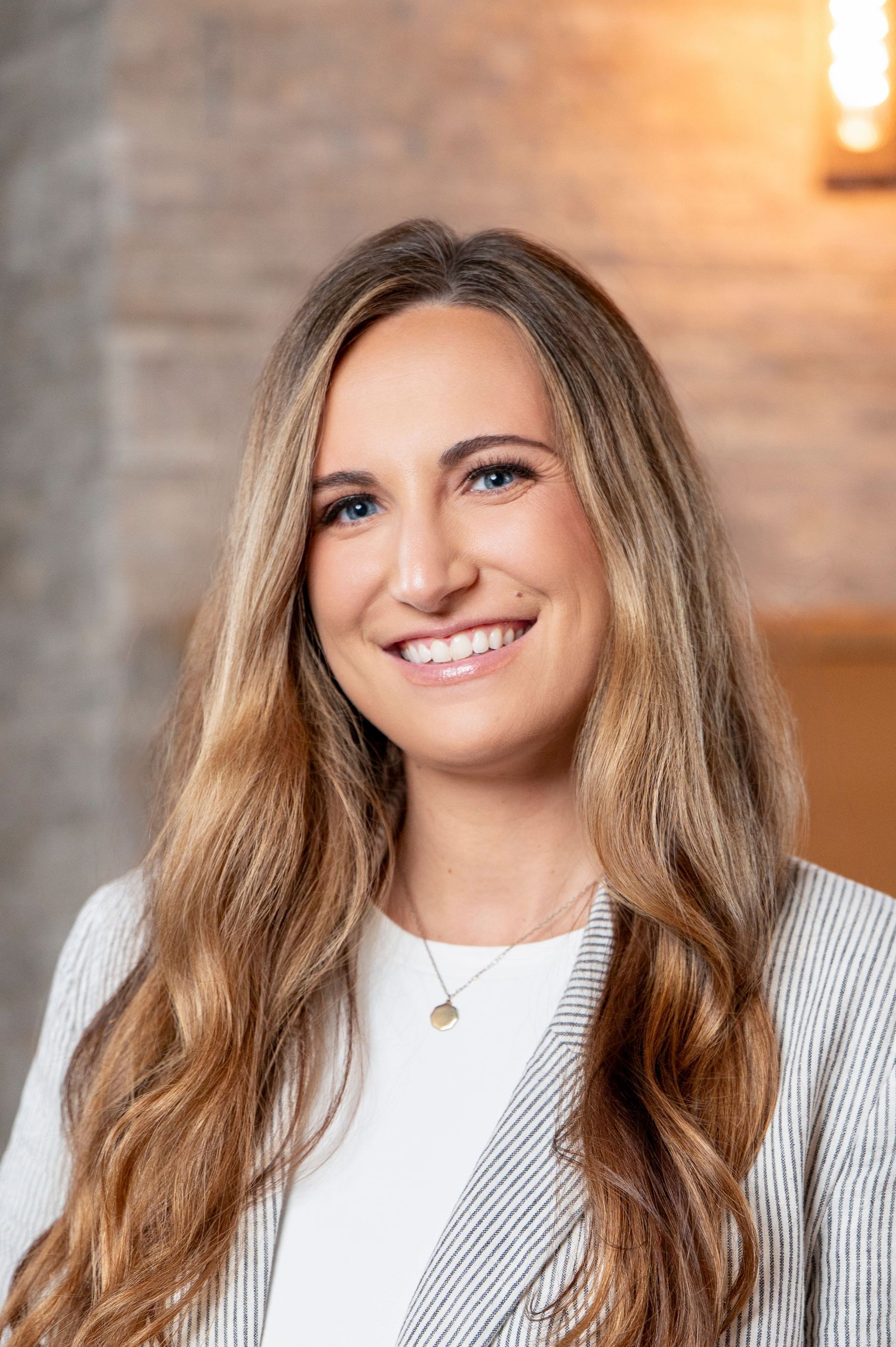 Smiling woman with long wavy hair in a white top and gray cardigan against a warm brick background