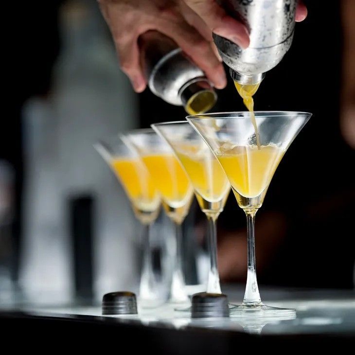 Bartender pouring yellow cocktail from shaker into martini glasses.