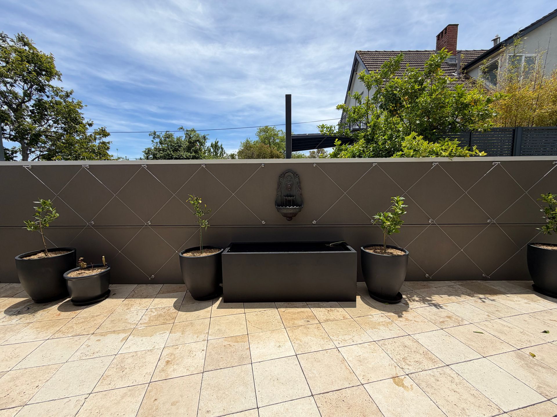 A dark rectangular water fountain and potted plants centered against a dark brown, diamond-patterned wall on a patio.  — Straight up Building Projects in Holt, ACT 