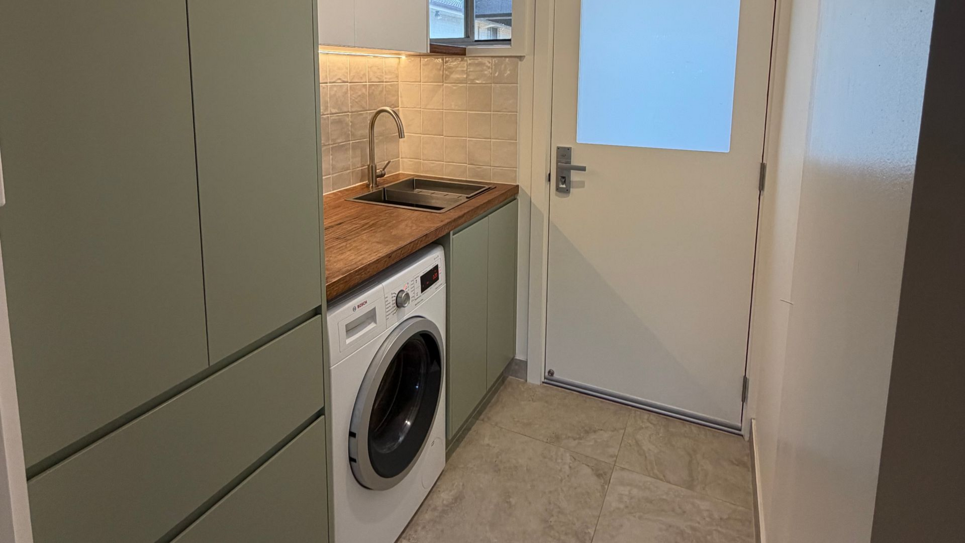 A small, modern utility room with sage green cabinets, a wood countertop, a washing machine, a sink, and a white door — Straight up Building Projects in Holt, ACT