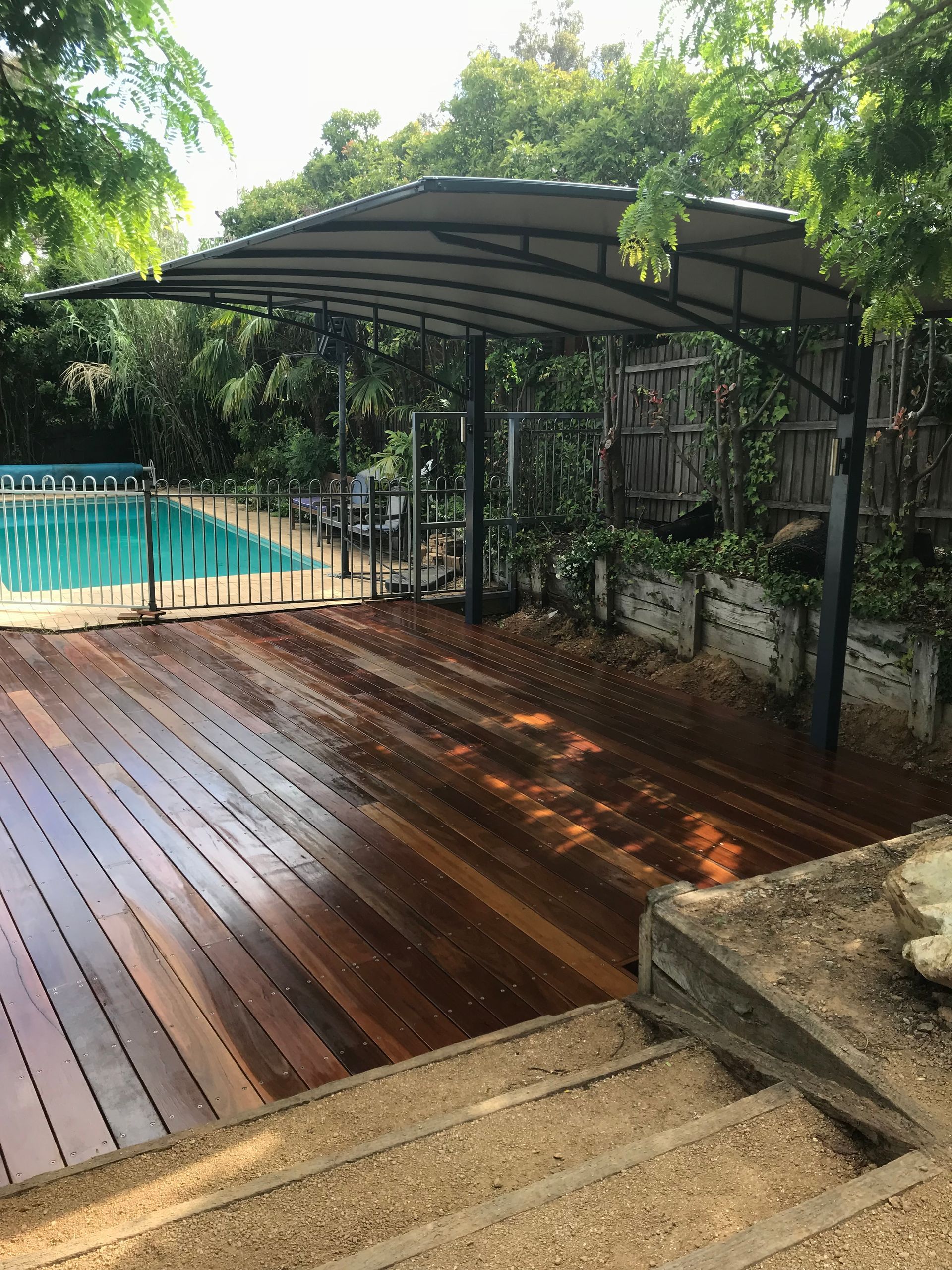 A wooden deck with a curved metal patio cover overlooks a swimming pool, set in a backyard with trees and a gravel path. — Straight up Building Projects in Holt, ACT