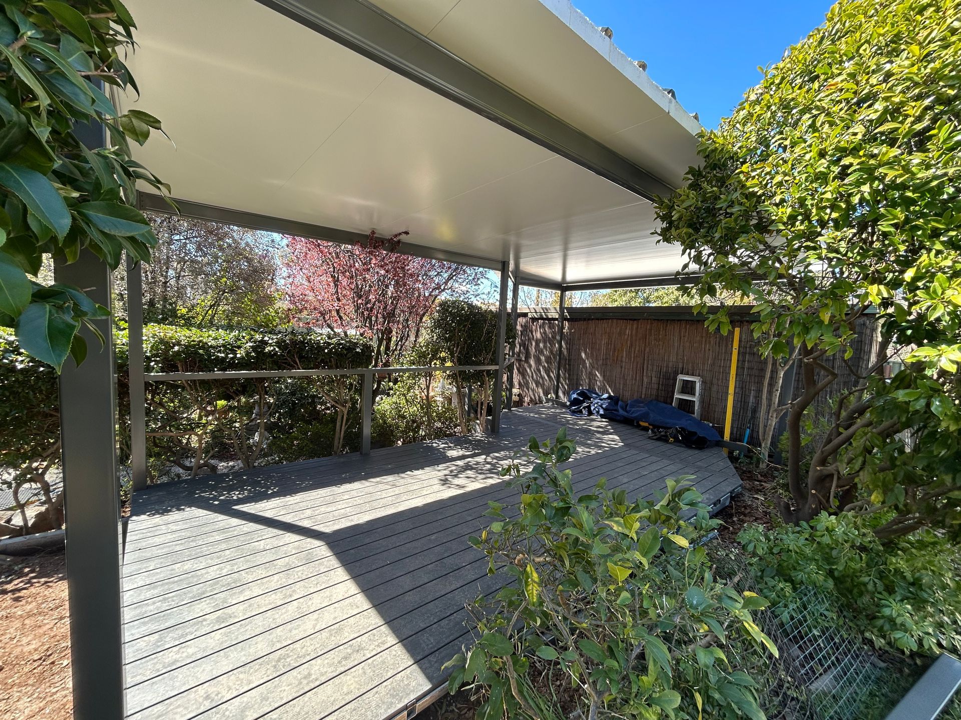 A covered wooden deck in a sunlit backyard, surrounded by lush green trees and foliage. — Straight up Building Projects in Holt, ACT