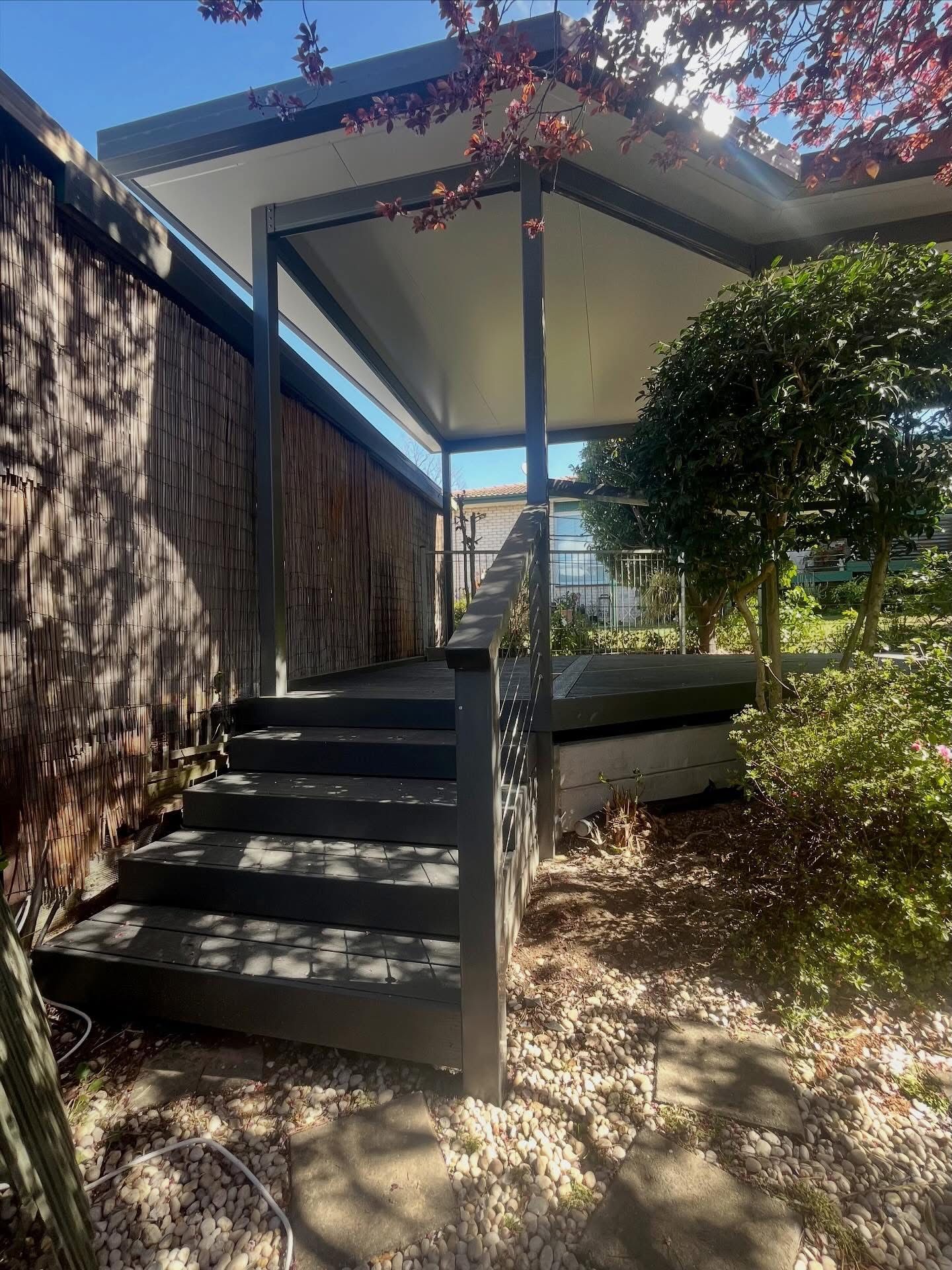 Dark grey wooden steps lead up to a covered outdoor deck area framed by a wooden fence and garden foliage  — Straight up Building Projects in Holt, ACT