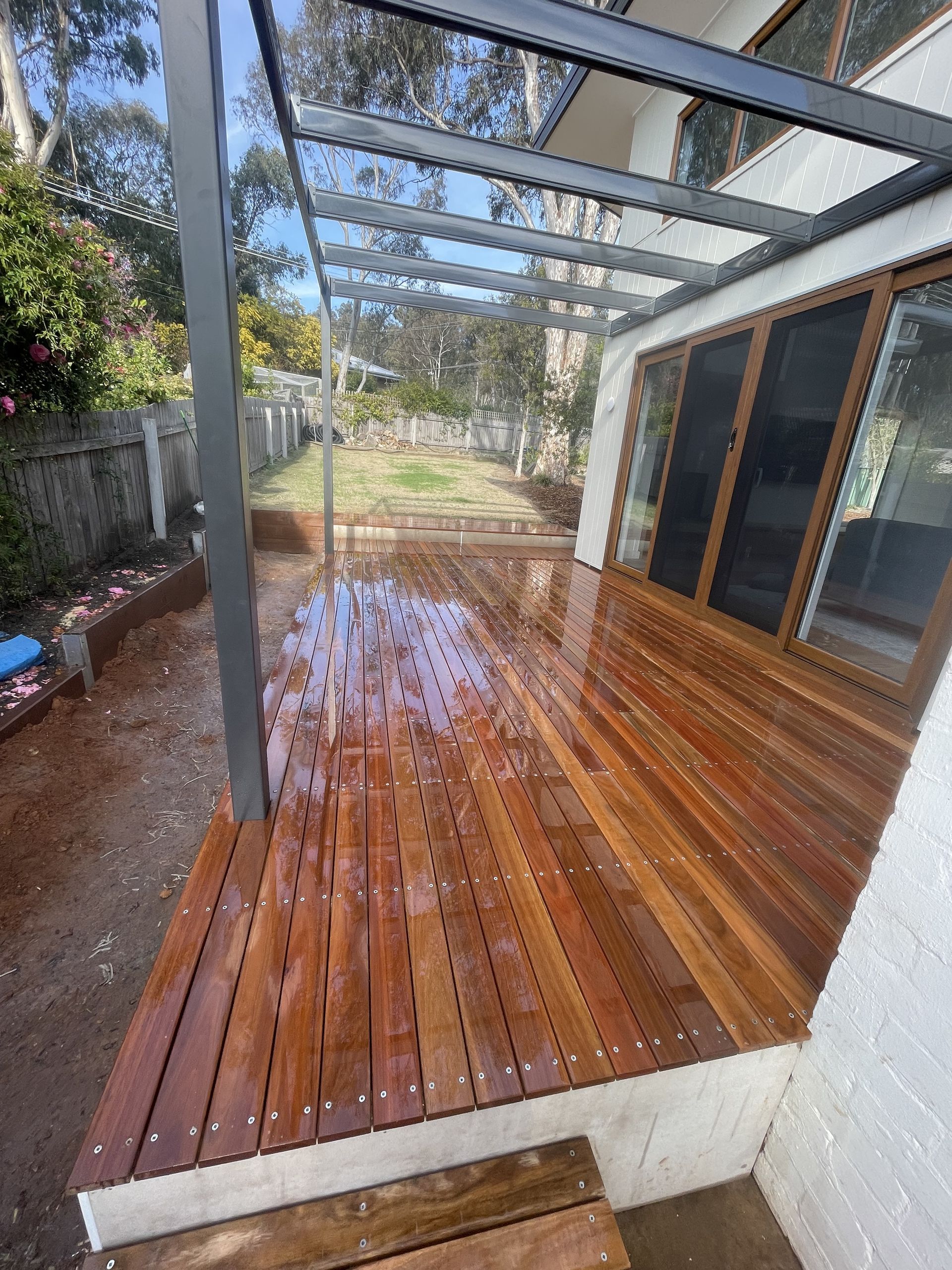 A newly stained wooden deck with a black steel pergola frame, adjacent to a house with sliding glass doors  — Straight up Building Projects in Belconnen, ACT
