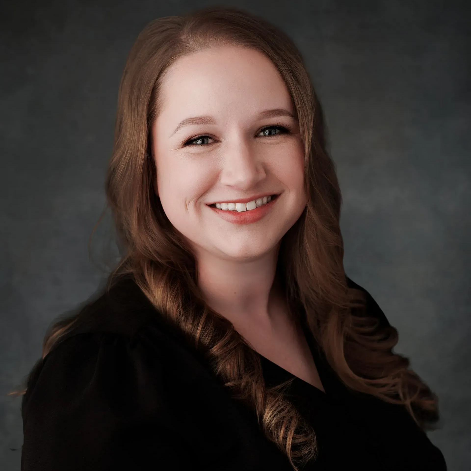 Woman with long brown hair smiles at the camera, wearing a black top. Blue-gray background.