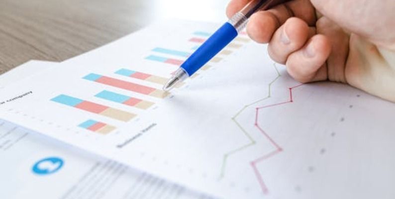 A hand holding a blue pen points to a bar graph and line graph on a printed page.
