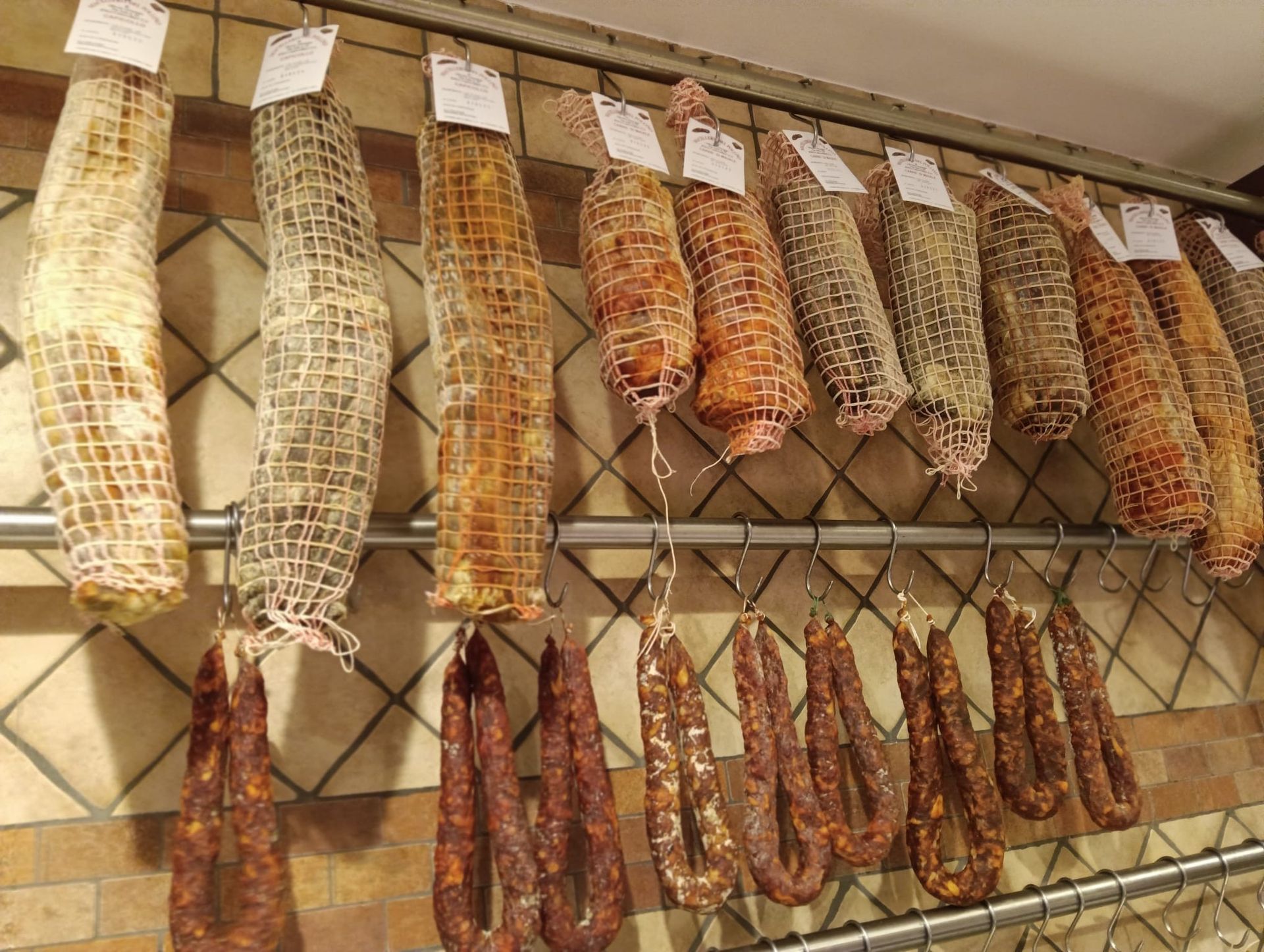 Vari salumi appesi su una rastrelliera di metallo contro una parete piastrellata.