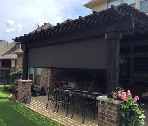 Patio with retractable shade, outdoor dining table, brick pillars, and potted pink flowers beside a house