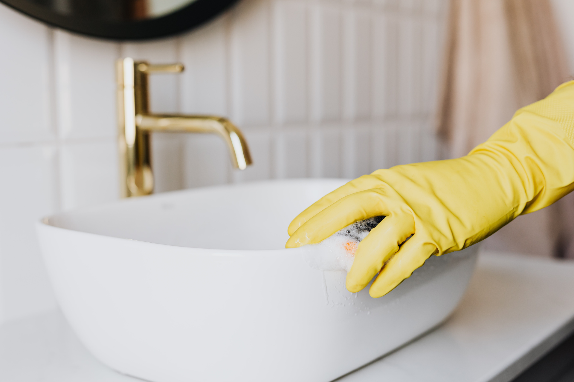 Gloved hand cleaning a white bathroom sink under a gold faucet