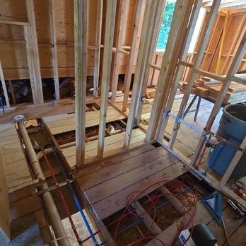 Interior view of a bathroom under construction with exposed wood framing, flooring, and plumbing.