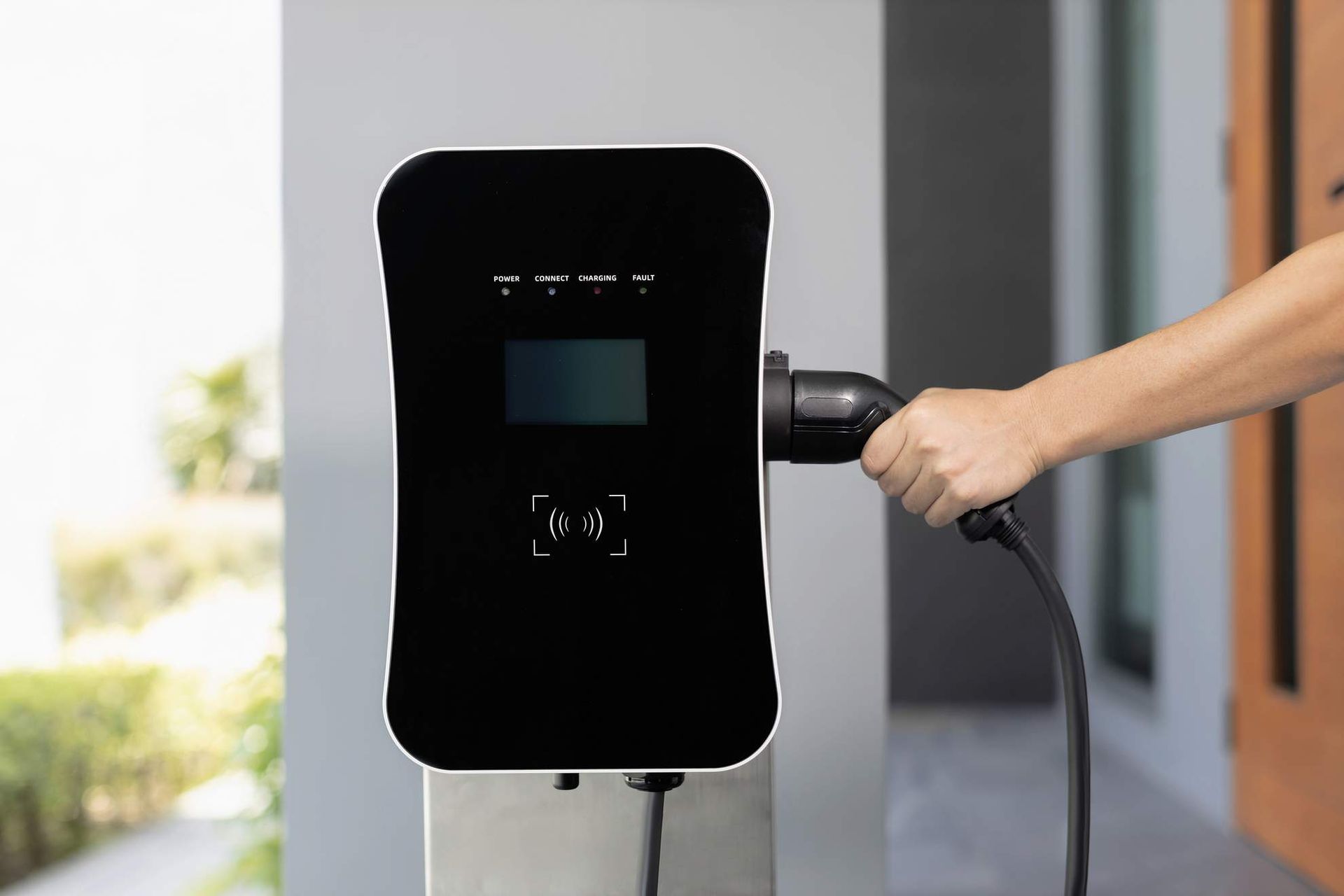 Persoon sluit de laadkabel van een elektrisch voertuig aan op een zwart laadstation buiten.