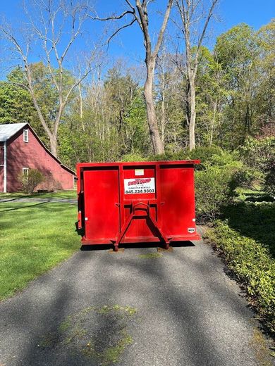 Red dumpster blocking a driveway beside a red barn in a wooded yard.