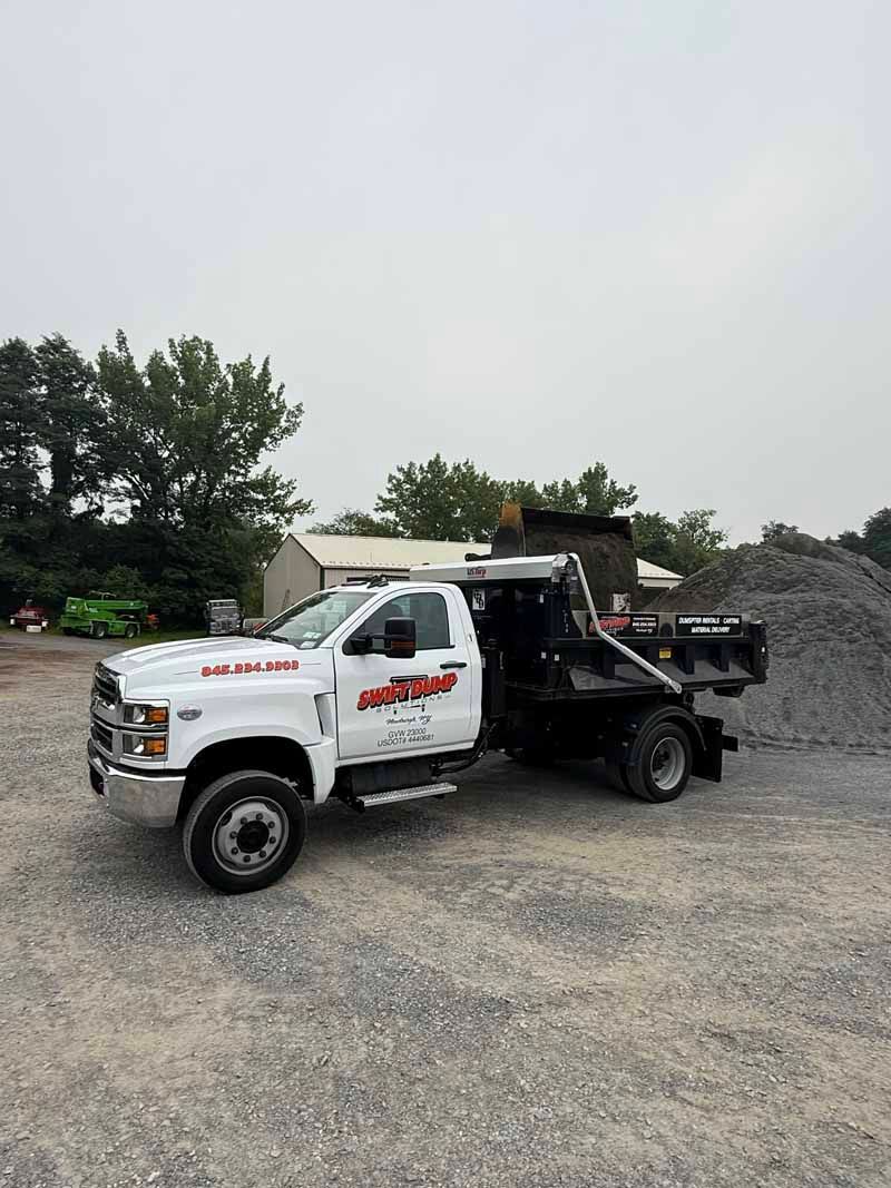 White tow truck with boom parked on gravel beside a pile of dirt under an overcast sky.