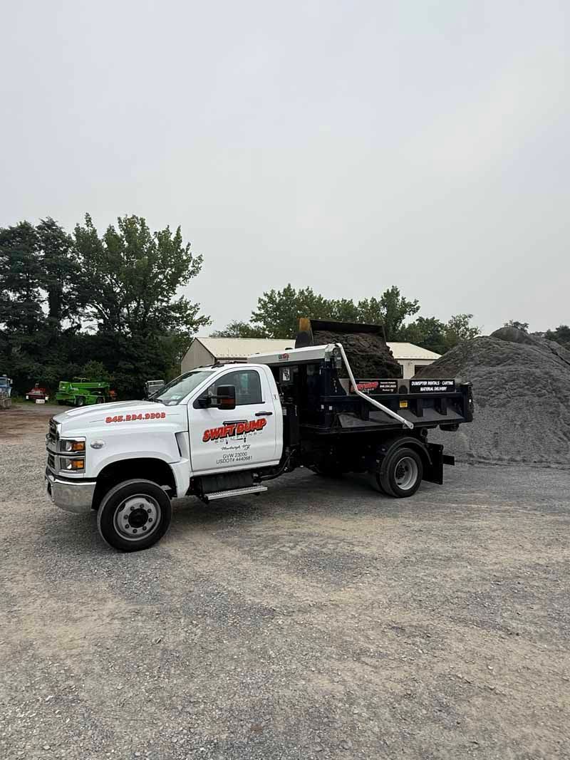 White dump truck parked on gravel near a pile of gray rock under an overcast sky