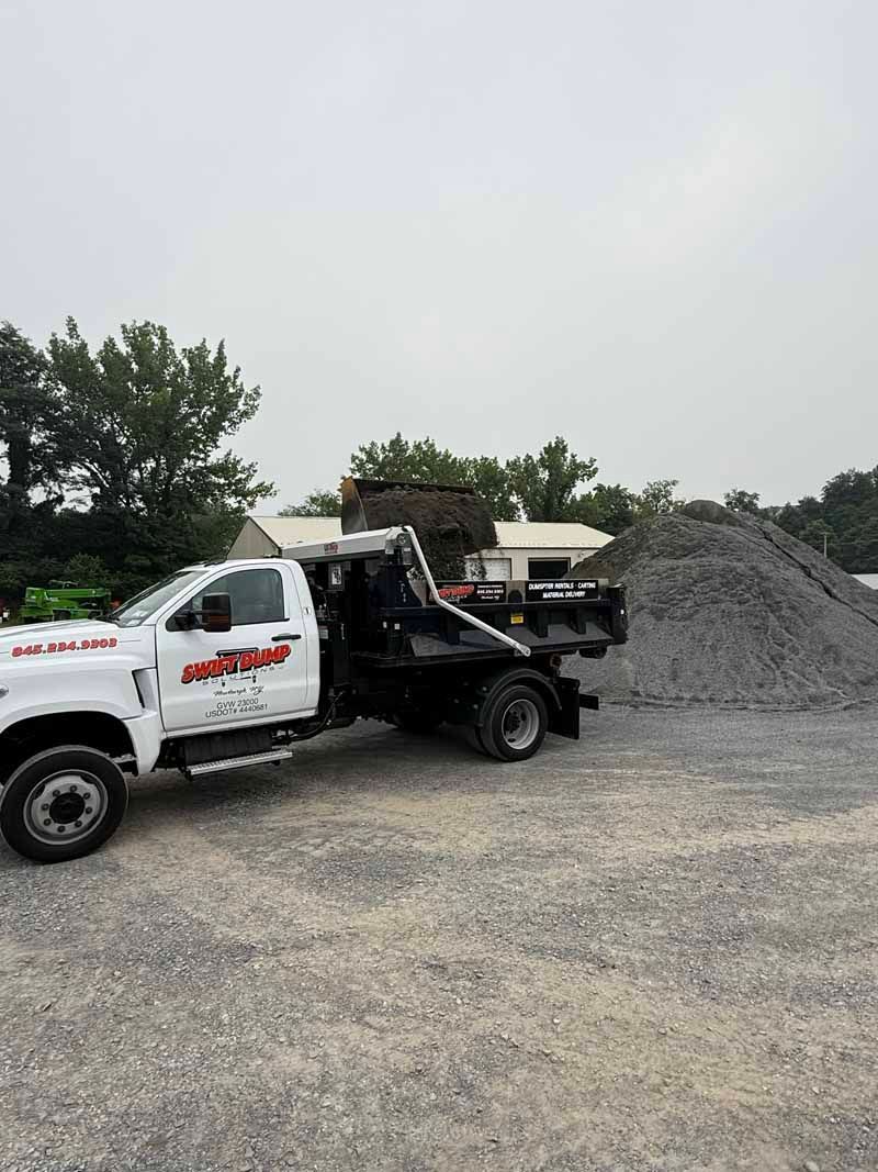 White dump truck unloading gravel beside a large gravel pile in an open lot under a gray sky