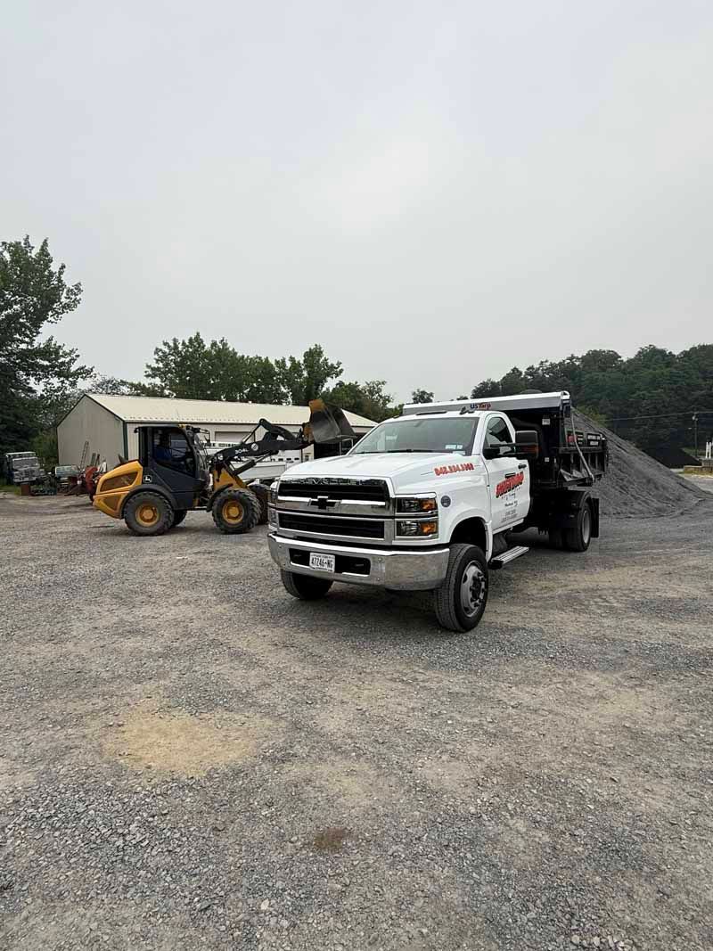 White pickup truck and loader on a gravel lot near a shed and trees under an overcast sky