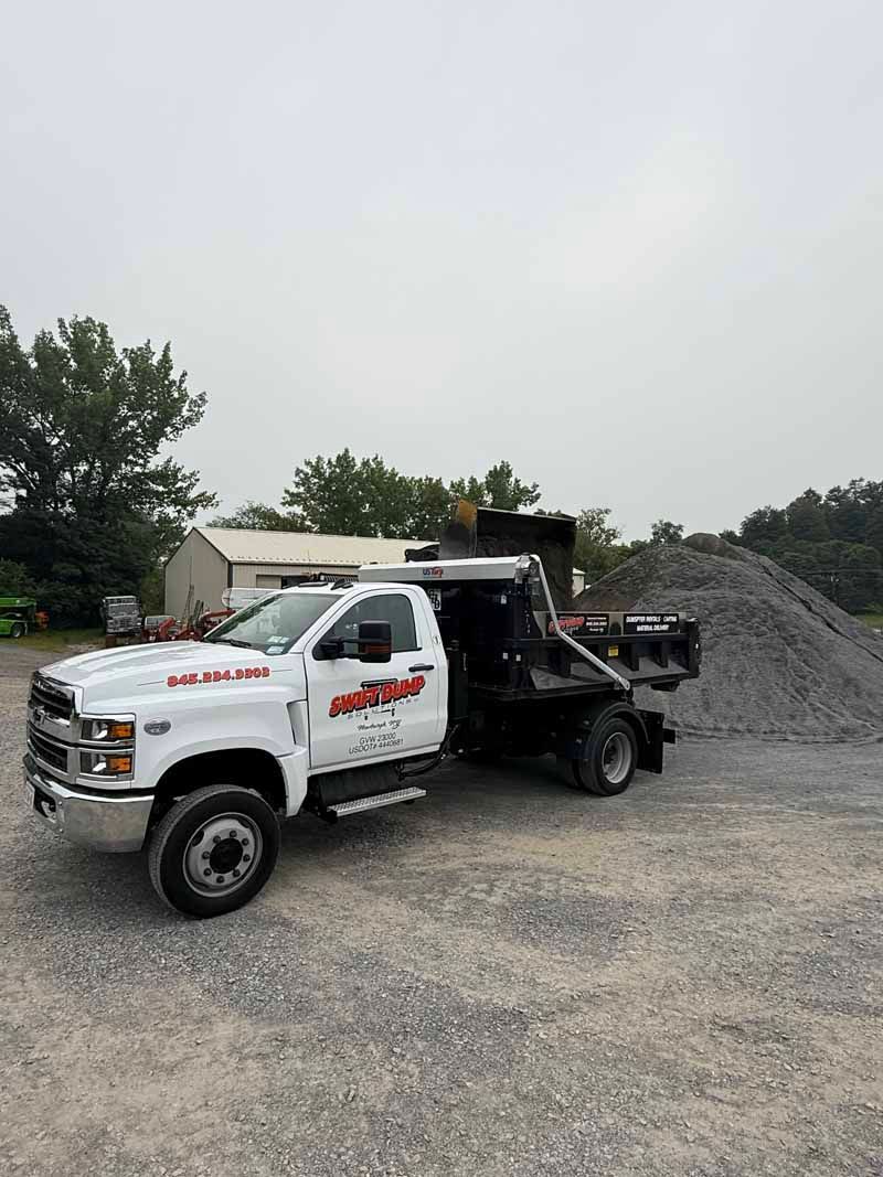 White dump truck with red logo parked on gravel beside a large pile of gravel outdoors