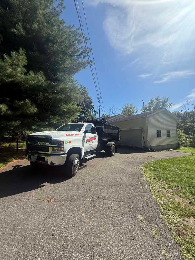 White utility truck and trailer parked on gravel driveway beside a small house, under a bright blue sky.