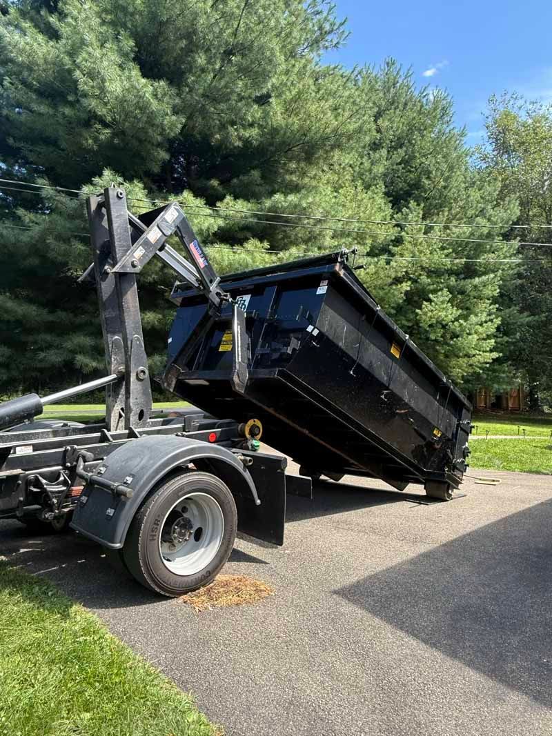 Black dump trailer raised on a truck chassis beside a gravel driveway and trees