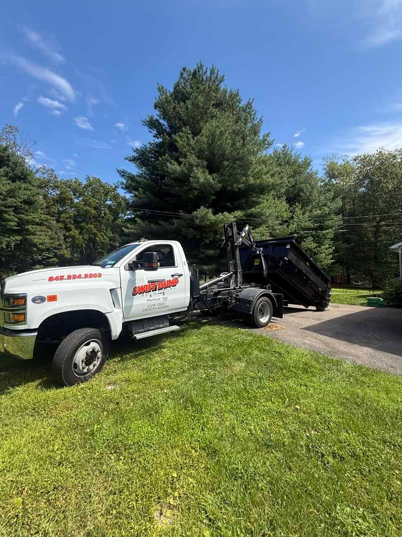 White dump truck with raised bed parked on grass beside a driveway under a blue sky.