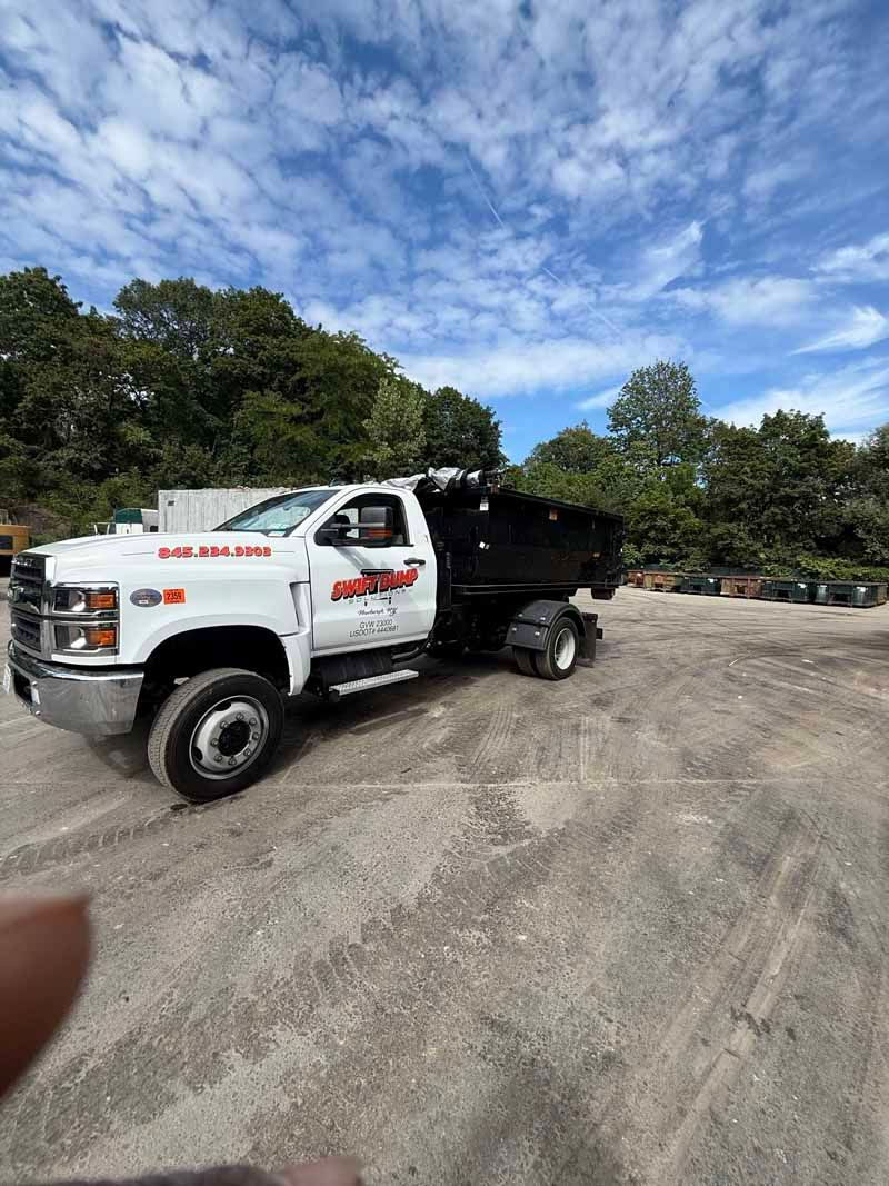 White dump truck parked on a gravel lot under a blue, cloudy sky, with trees in the background