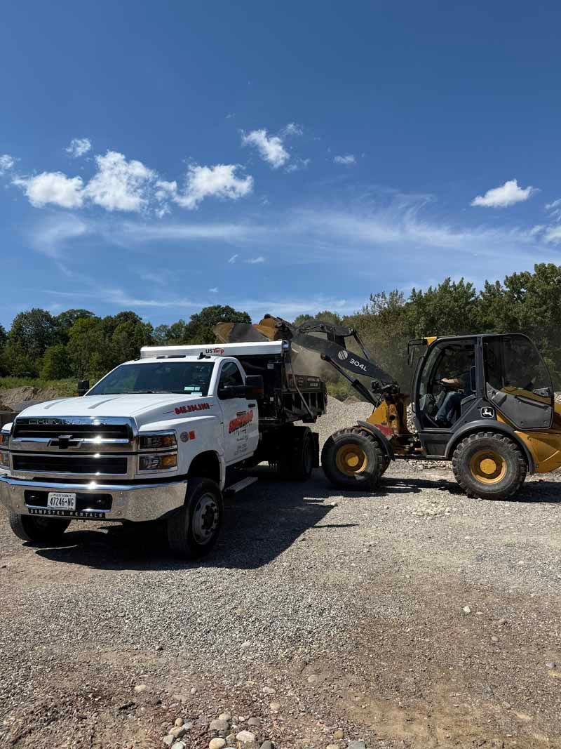 White tow truck and yellow backhoe loader parked on gravel under a blue sky.