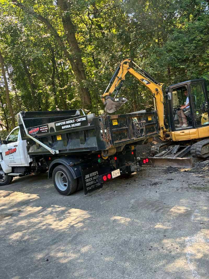 Yellow excavator loading debris into a black dump truck on a wooded roadside
