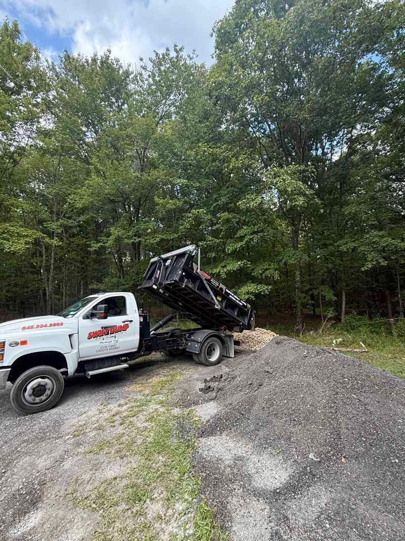 White dump truck with raised black bed parked on a gravel pile near a wooded area.