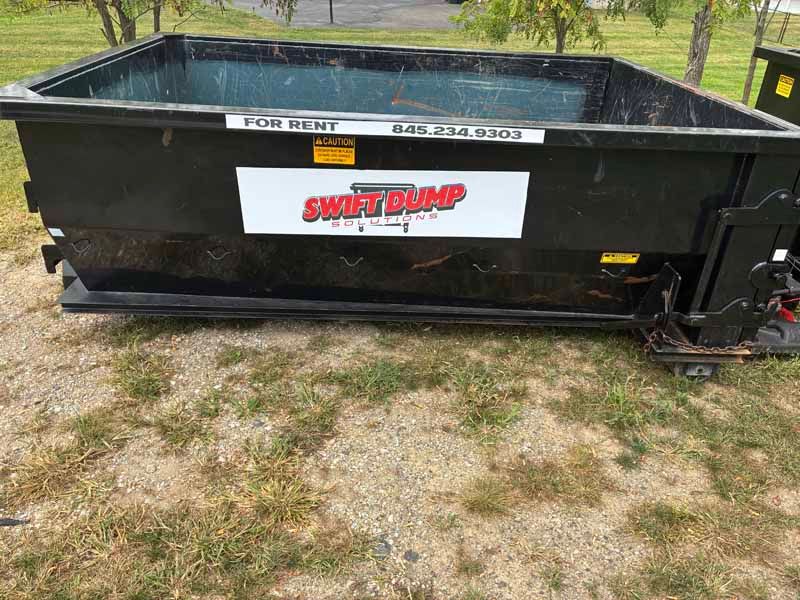 Black metal dump trailer on grass with red-and-white logo and warning labels.