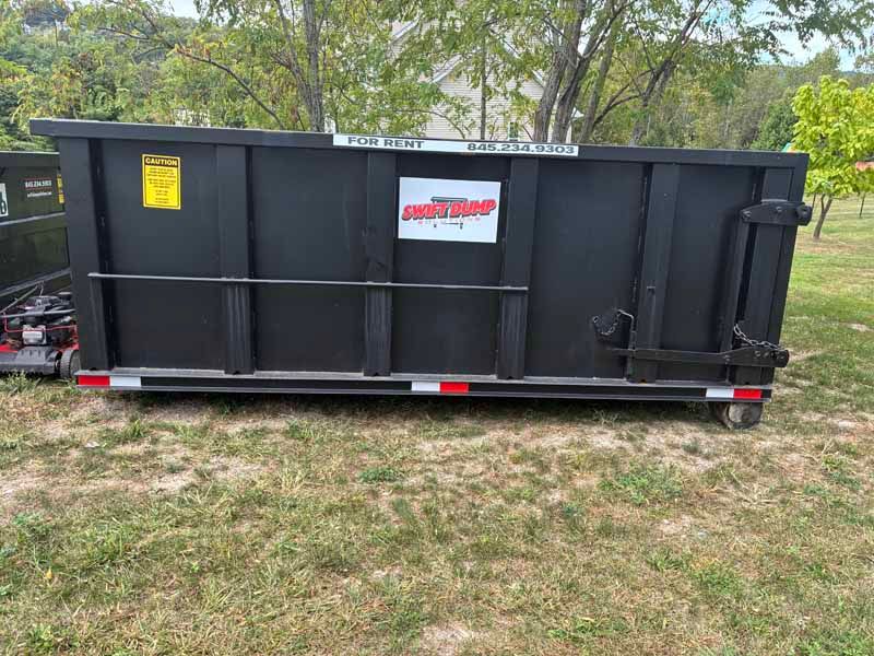 Large black dumpster trailer parked on grass with a white-and-red sign, trees in the background