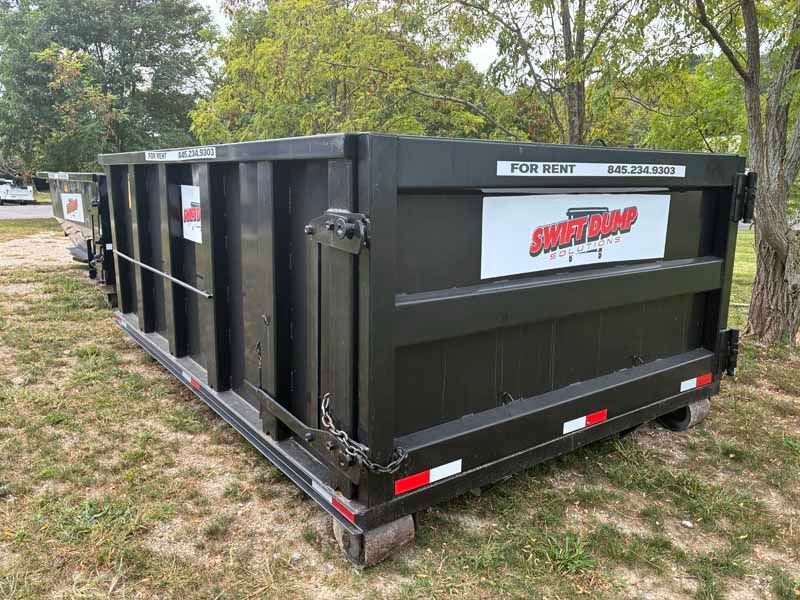 Black dumpster container with white and red logo, parked on grass beside a road.
