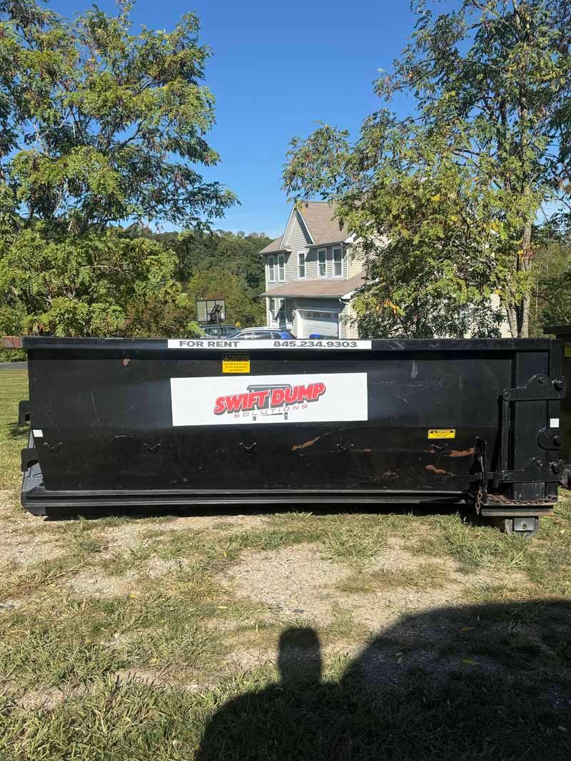 Black dumpster in a grassy yard with a house and trees in the background.