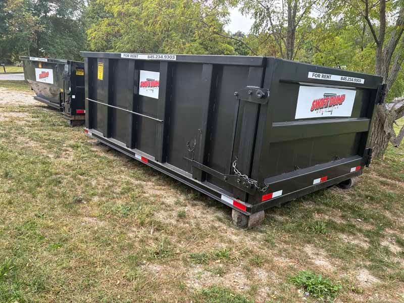 Large black roll-off dumpster on grass beside a wooded area, with white/red logo panels.