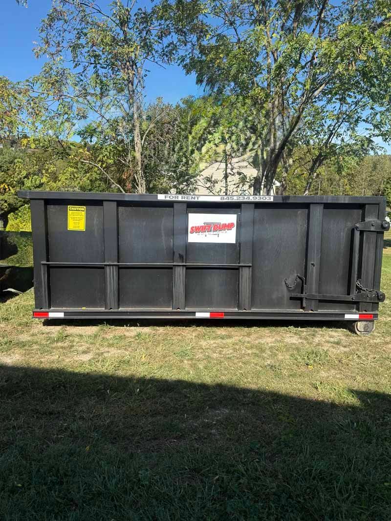 Black construction dumpster on grass with trees in the background