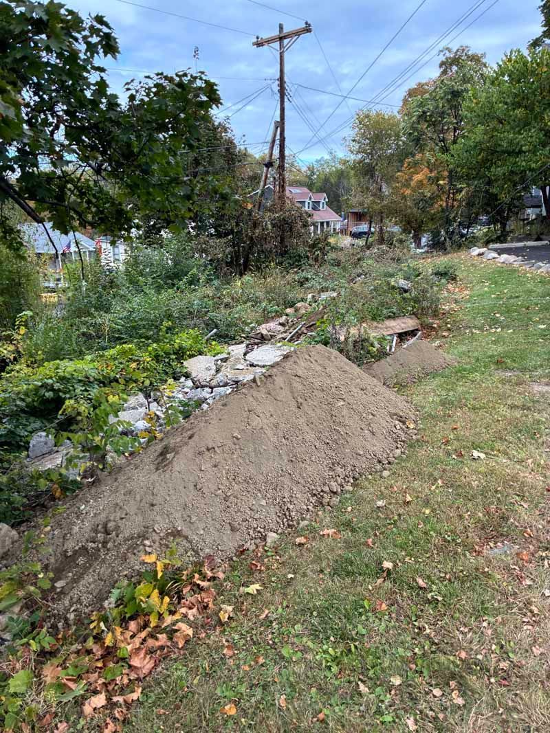 Dirt pile beside a grassy roadside ditch with trees, utility poles, and houses in the background