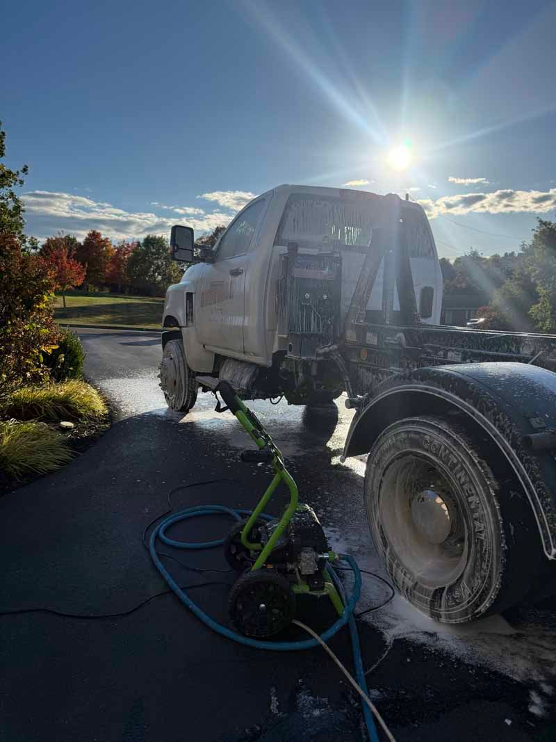 Truck being pressure washed in a driveway, with soapy foam and bright sun in the background.