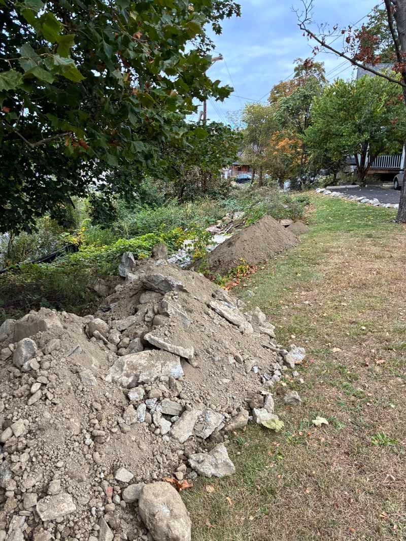 Dirt and rubble piles beside a grassy path with trees and shrubs in a park-like area