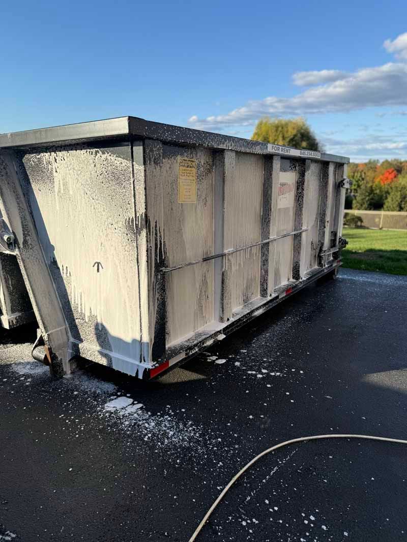 Large white dumpster on asphalt, with debris scattered around and a hose in the foreground.