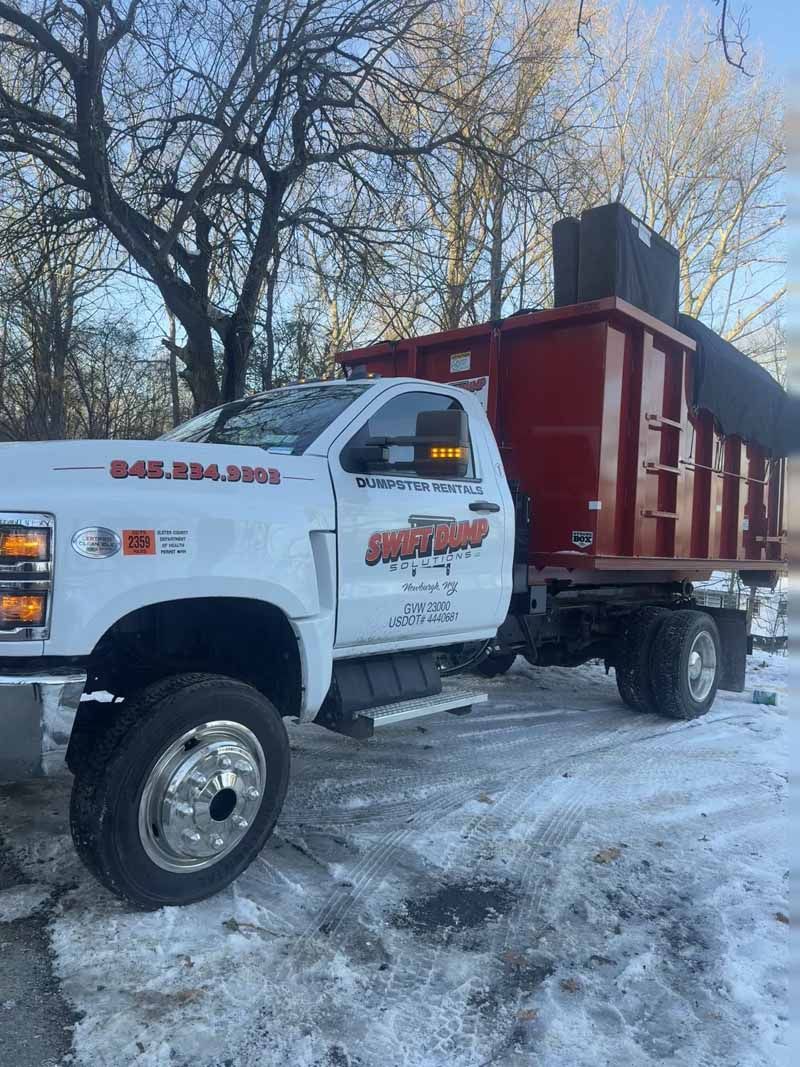 White dump truck with red bed parked on snowy ground near bare trees.