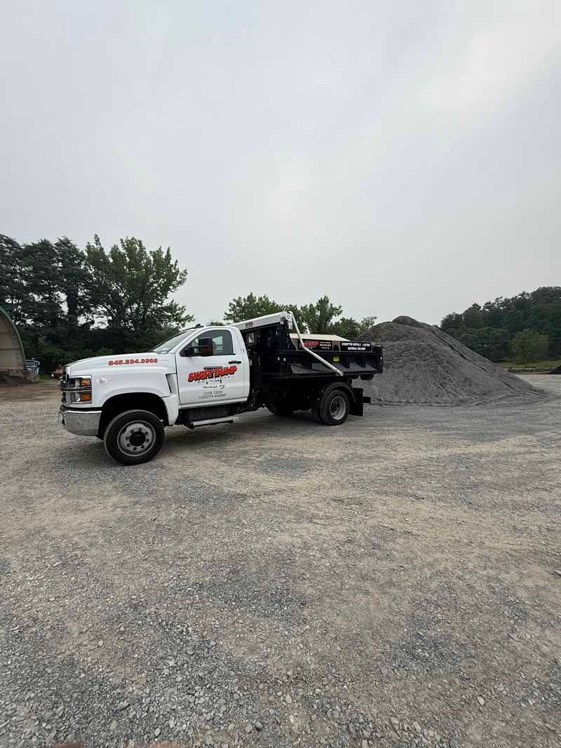 White tow truck parked on a gravel lot beside a black car carrier and a pile of gravel under an overcast sky