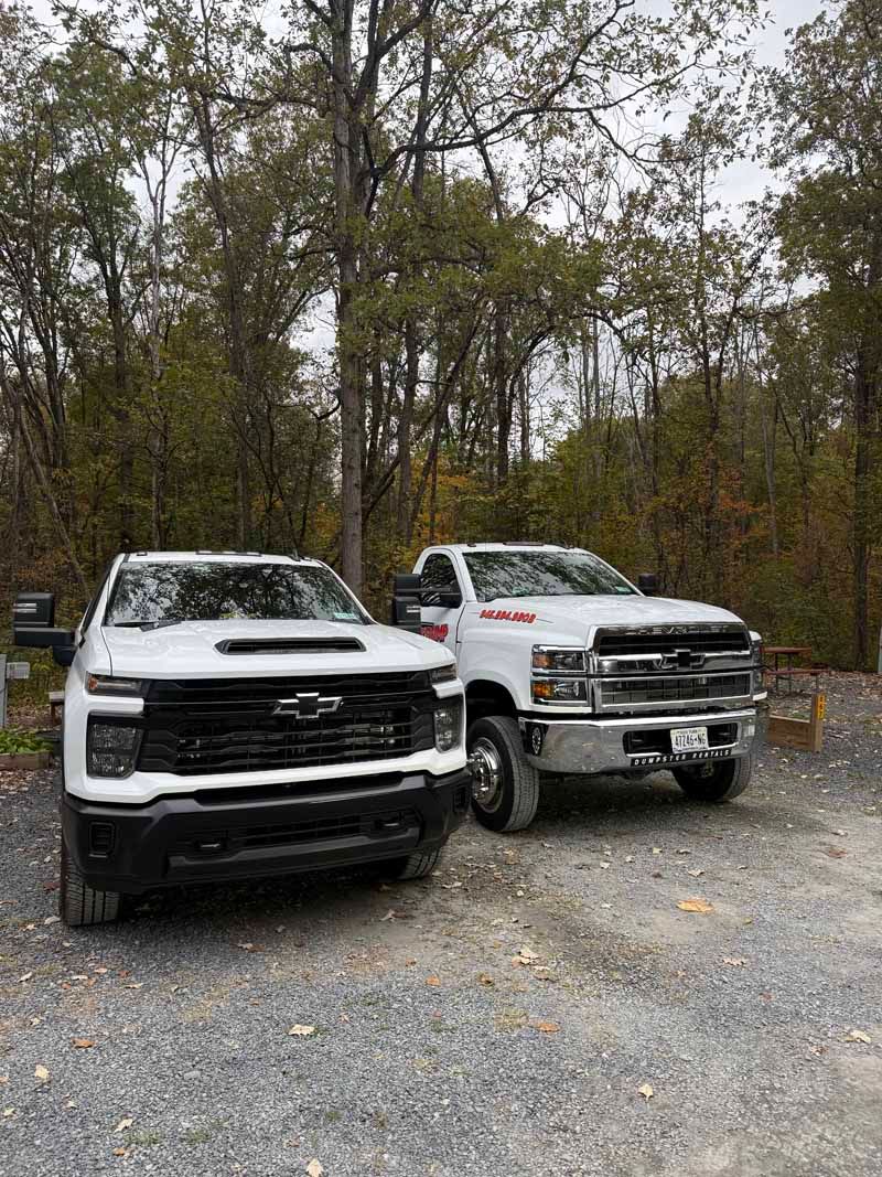 Two white pickup trucks parked on a gravel road in a wooded area with autumn trees