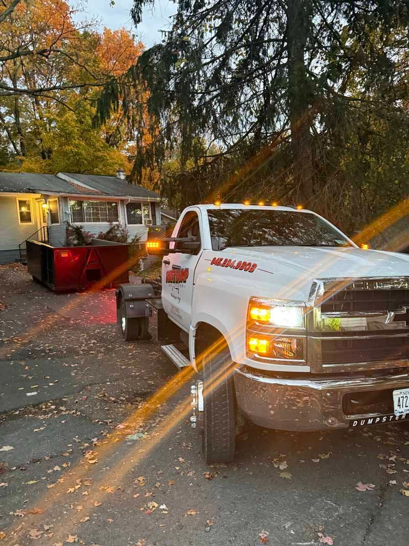 White utility truck and trailer parked on a leaf-covered street in autumn, with headlights on at dusk